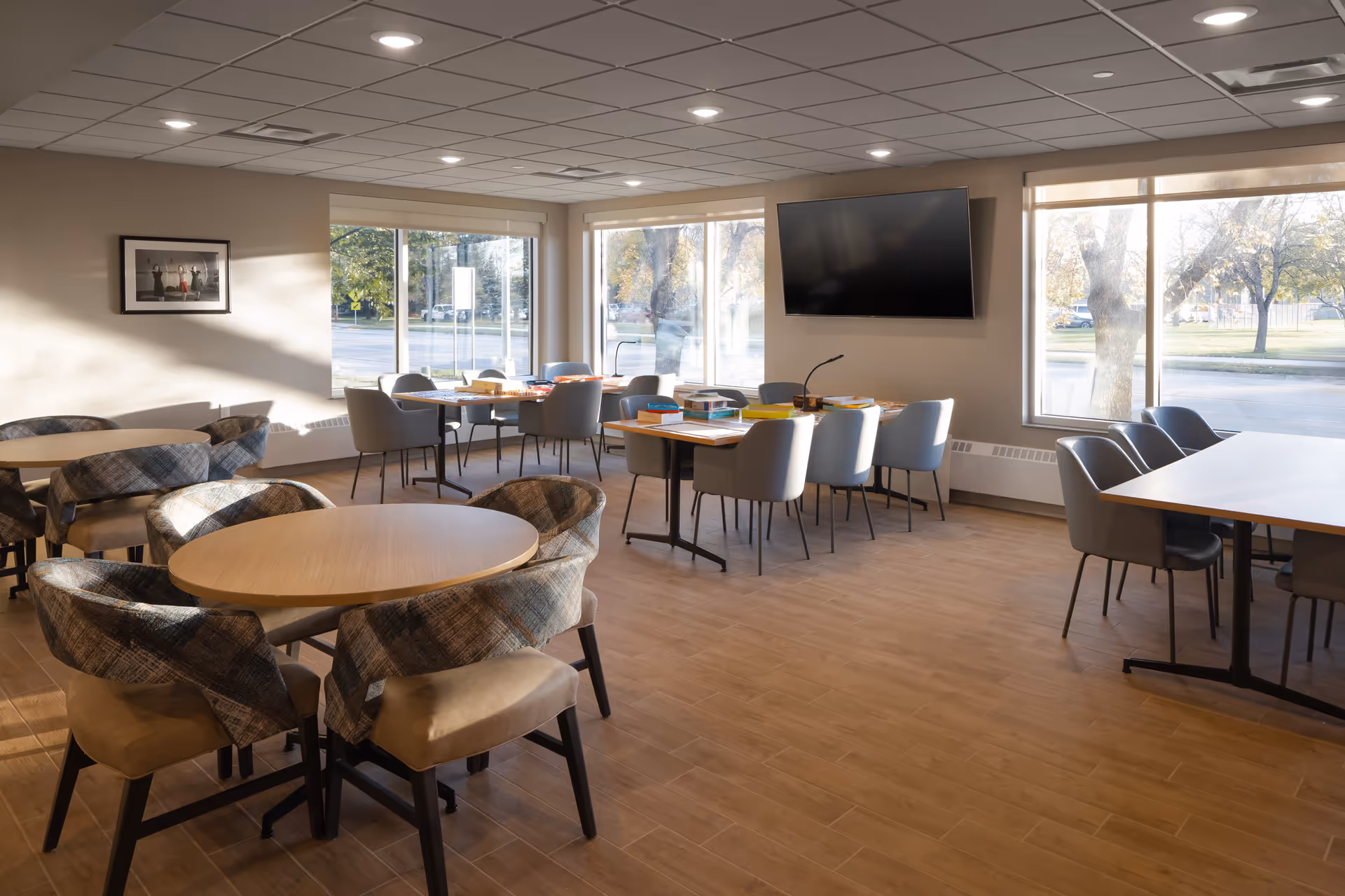 A bright and spacious common area with multiple tables and chairs arranged for group activities or dining. Large windows allow natural light to fill the room, and a flat-screen TV is mounted on the wall. The room has a light wood floor and a ceiling with recessed lighting.