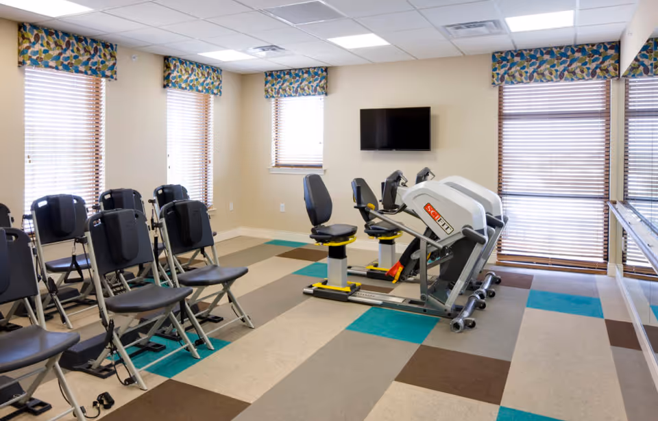 A bright interior activity room with exercise machines, rows of folding chairs, a wall-mounted TV, and patterned window valances.