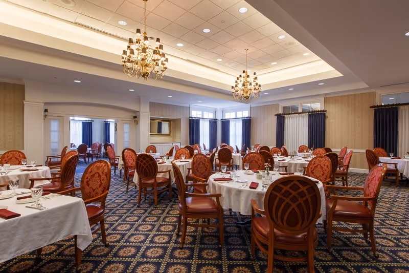Spacious formal dining room with round tables covered in white linens and red upholstered chairs beneath chandeliers.