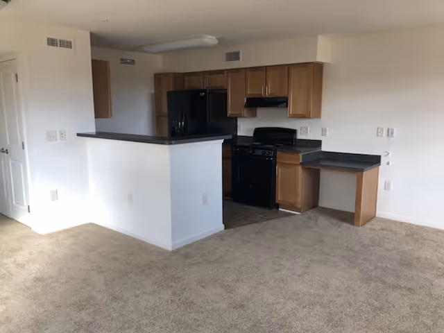 Interior view of a small kitchen area in an apartment with wooden cabinets, a black refrigerator, black stove, and a small built-in desk area. The kitchen is partially enclosed by a half wall with a dark countertop, and the surrounding floor is carpeted in beige.