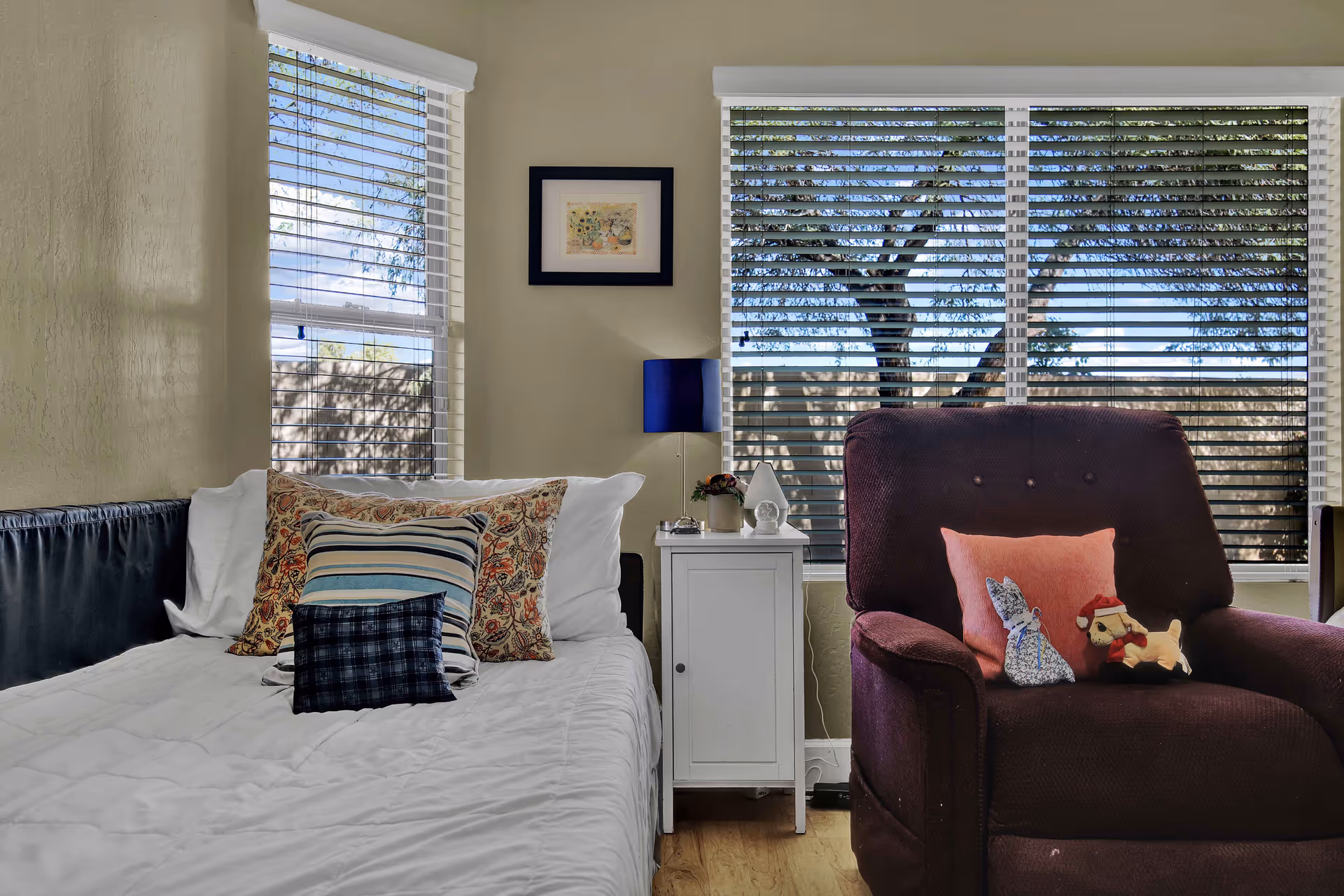 A cozy bedroom with a neatly made bed featuring multiple decorative pillows, a white nightstand with a blue lamp and small decor items, and a dark purple armchair with two stuffed animals and a pink pillow. Large windows with white blinds allow natural light to fill the room and show a view of trees outside.