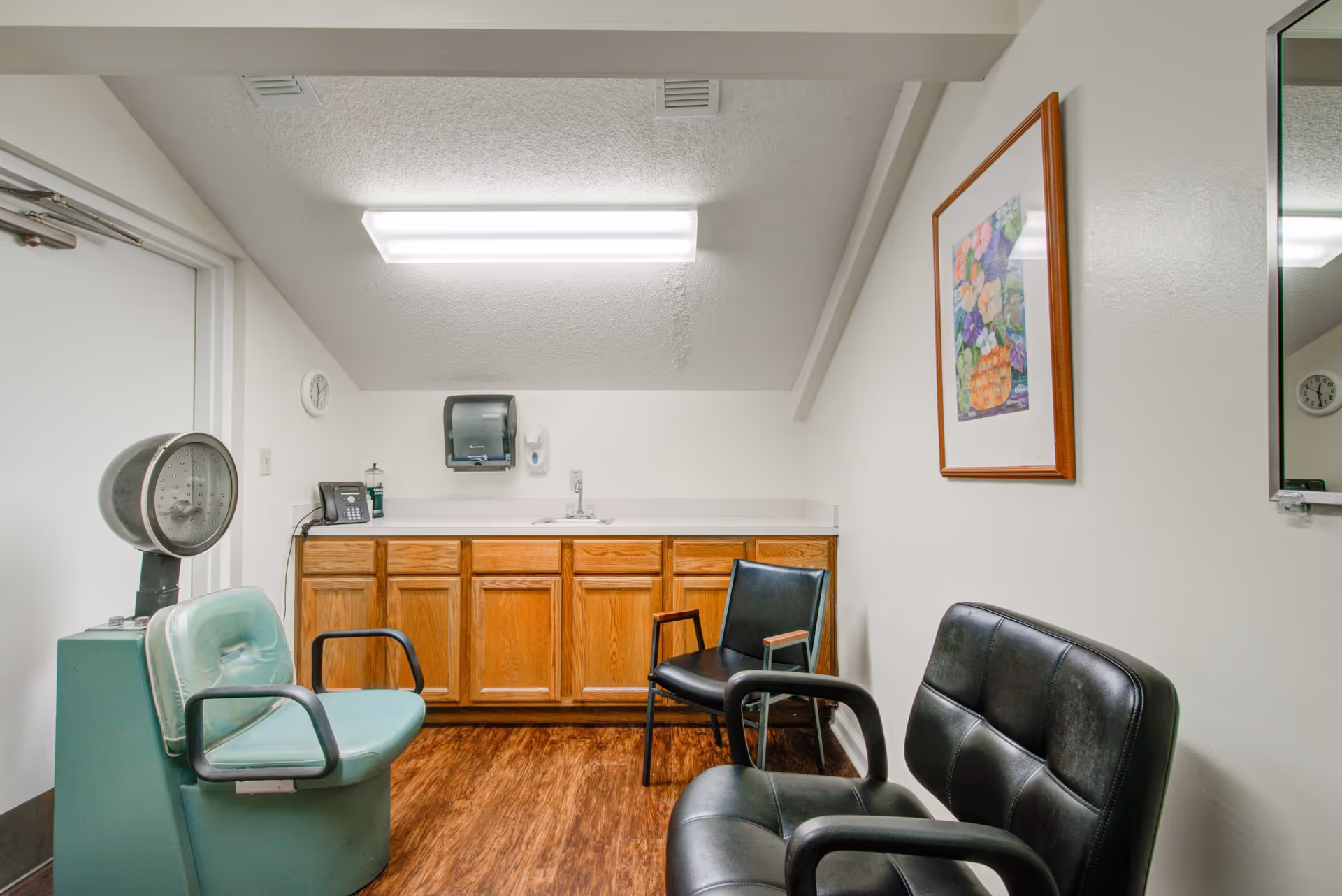 A small room with a wooden floor and white walls, featuring a green vintage medical scale chair, two black leather chairs, a wooden cabinet with a sink, a wall-mounted paper towel dispenser, a soap dispenser, a telephone, a clock, and a framed floral picture on the wall.