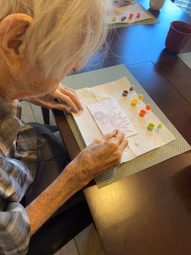 An elderly person with white hair is seated at a table, engaging in a painting activity. The person is holding a paintbrush and working on a small coloring sheet with a floral design. Several small paint pots with different colors are arranged on a paper towel on the table. The setting appears to be a communal or activity area with placemats and cups on the table.