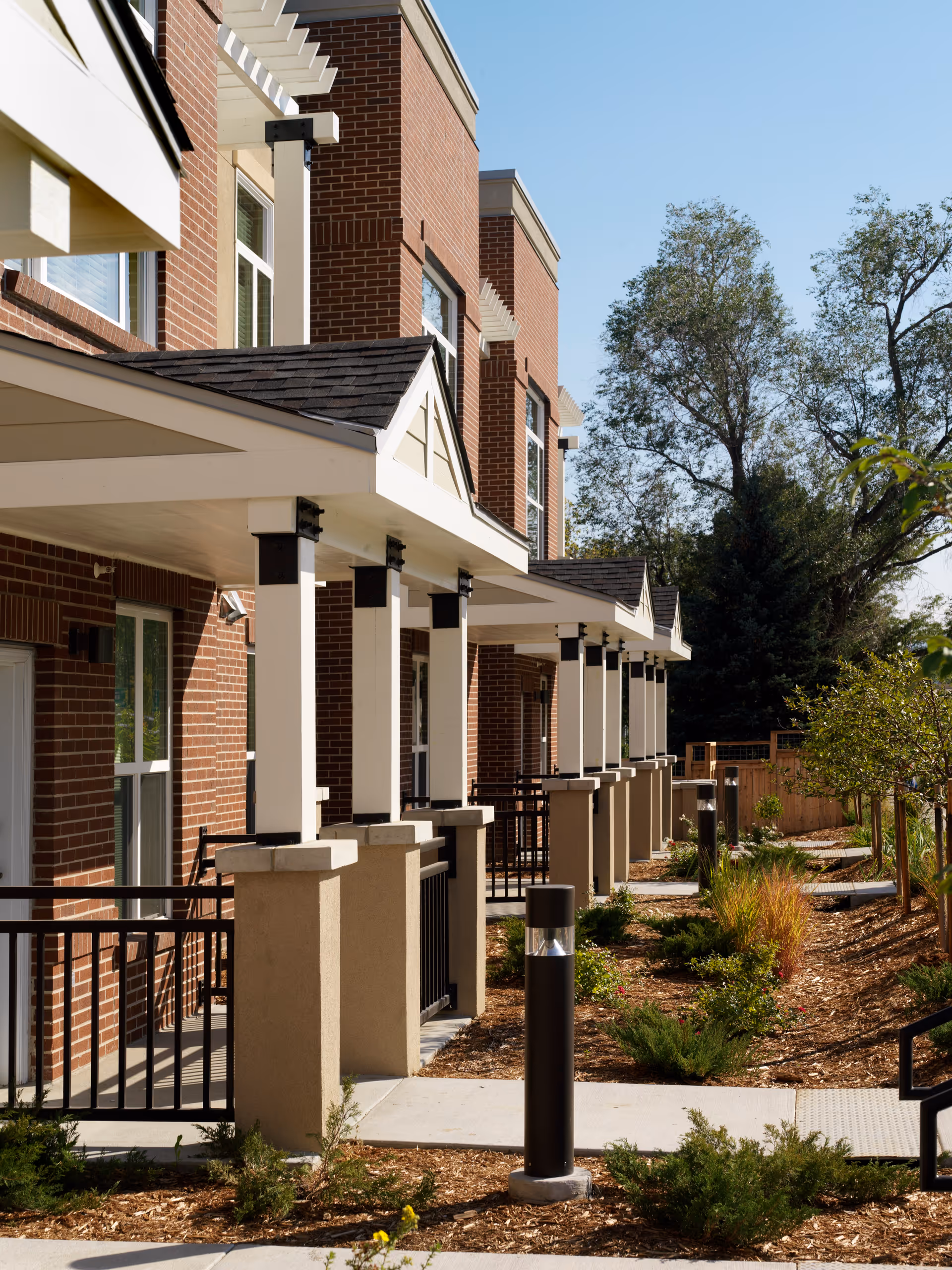 Exterior view of a senior living facility showing a row of brick buildings with covered porches, white columns, black railings, and landscaped garden beds along a paved walkway under a clear blue sky.