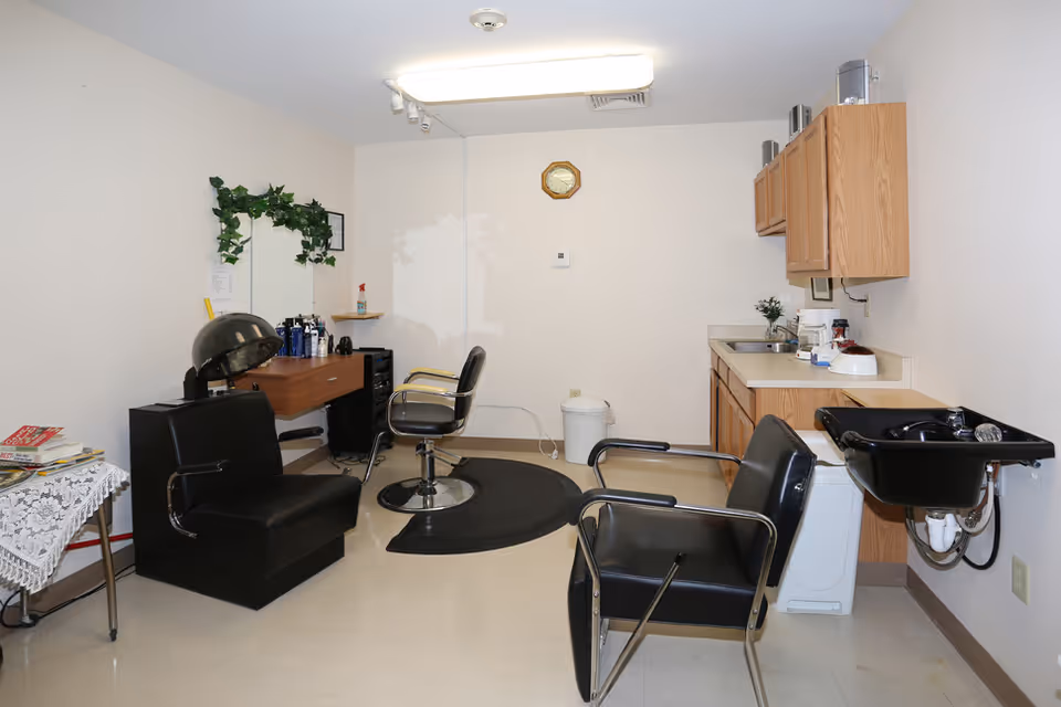 Interior view of a small hair salon area in a senior living facility with black salon chairs, a hair dryer, a mirror with green decorative leaves above it, a small table with magazines, wooden cabinets, a sink, and various hair care products on the counter.
