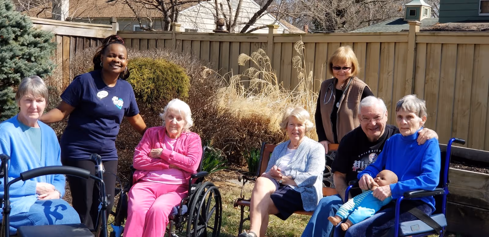 A group of elderly people and two caregivers are gathered outdoors in a garden area. Some elderly individuals are seated in wheelchairs, while others sit on benches. One elderly woman holds a baby doll. The caregivers are standing behind them, smiling. The background shows a wooden fence, bushes, and trees.