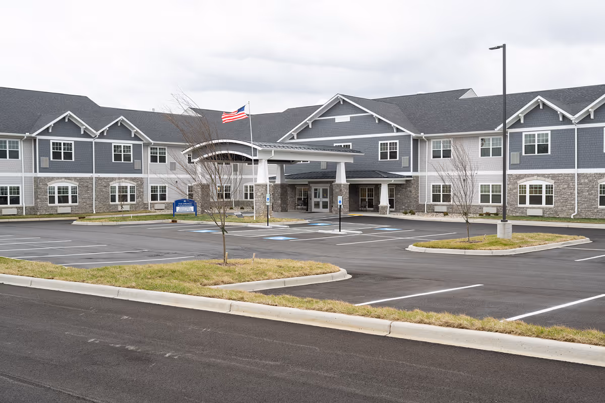 Front exterior of a two-story senior living facility with a covered entrance, empty parking lot, and an American flag.