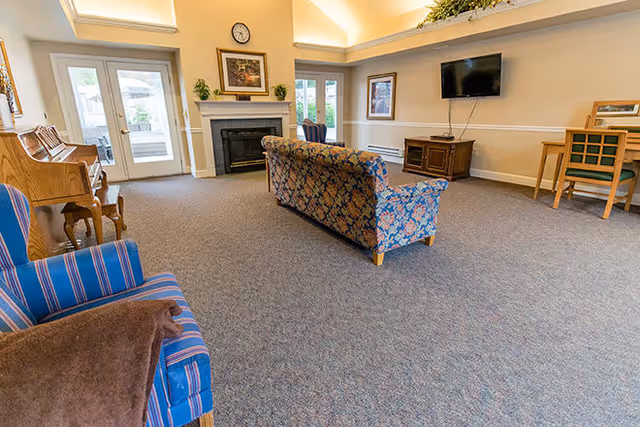 A spacious living room with a patterned sofa, a blue striped armchair with a brown blanket, a wooden piano, a fireplace with a clock and framed pictures above it, a flat-screen TV mounted on the wall, and a small desk with a chair. The room has carpeted flooring and large windows letting in natural light.
