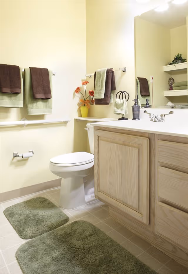 A clean bathroom with a toilet, wooden vanity with a sink, large mirror, green and brown towels hanging on the wall, green bath mats on the floor, and a small yellow vase with orange flowers on the countertop.