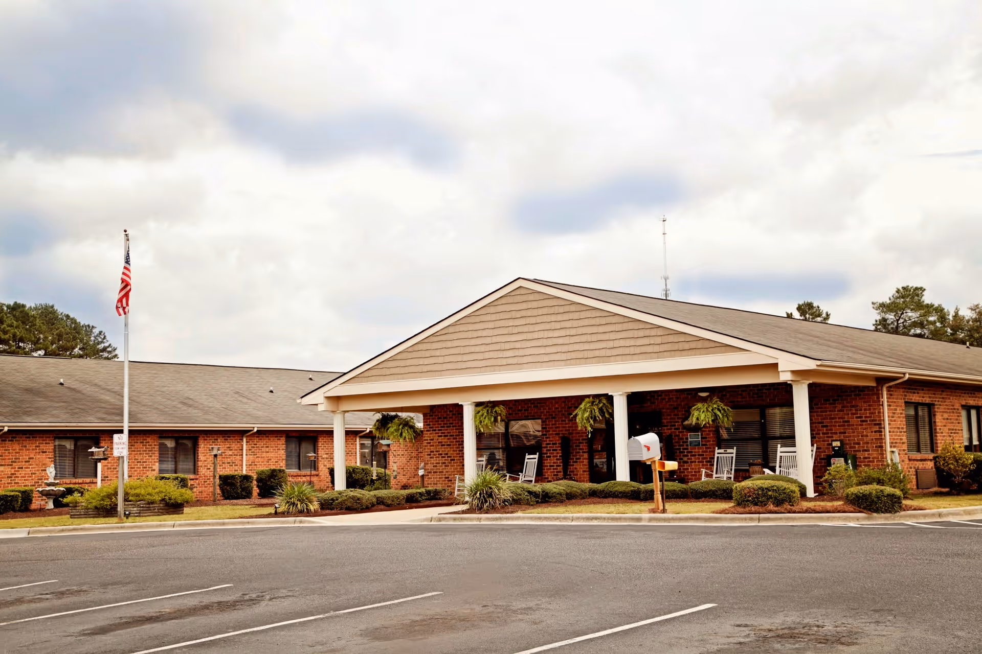Single-story brick senior living facility front entrance with a covered portico, rocking chairs, an American flag, and a parking lot.