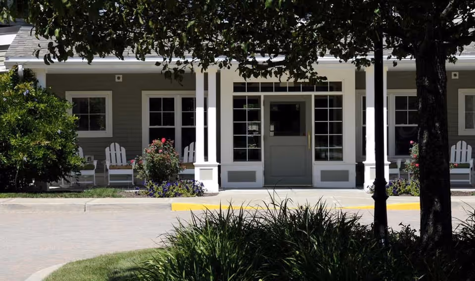 Front exterior view of a building with a covered porch featuring white columns, several white Adirondack chairs, and flower beds with blooming plants. Trees and shrubs partially shade the area in front of the entrance.