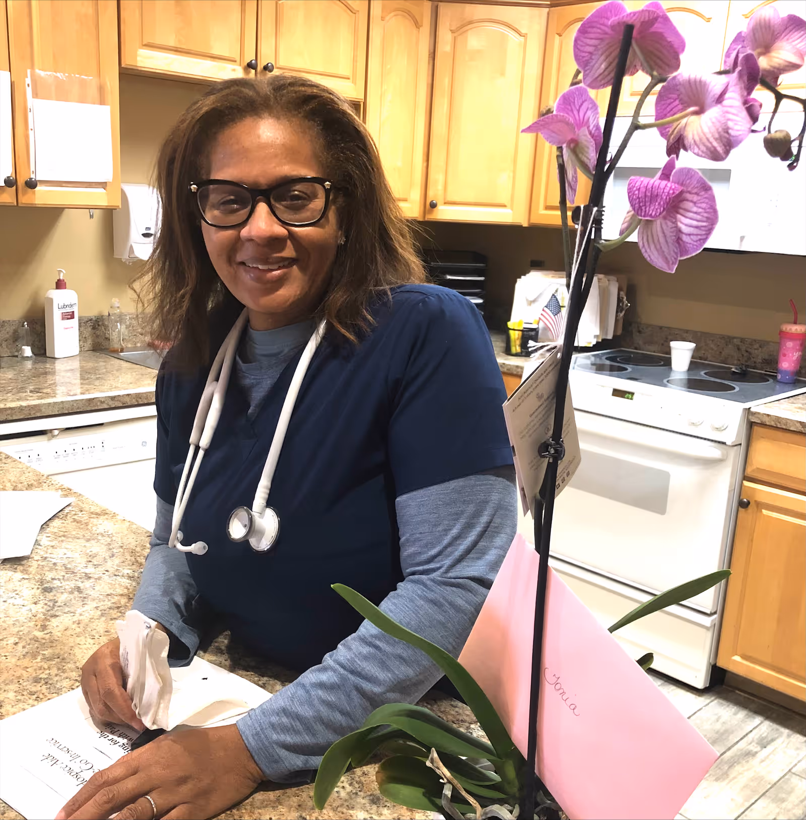 A smiling healthcare worker wearing glasses and a stethoscope around her neck stands behind a kitchen counter in a facility kitchen area. There is a pink orchid plant with a card and envelope on the counter, and kitchen cabinets, a stove, and a dishwasher are visible in the background.