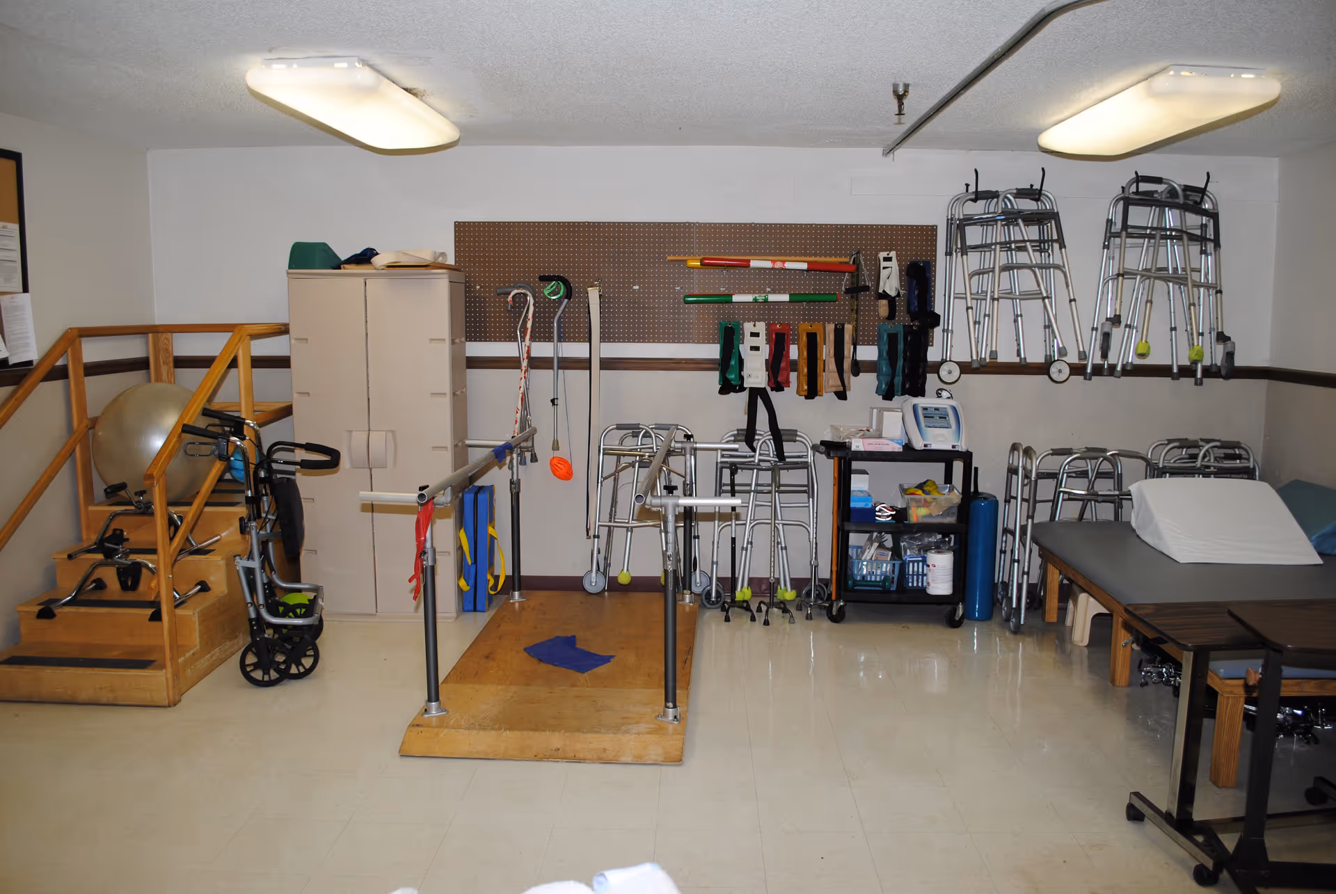 A therapy room with parallel bars, walkers, exercise equipment and therapy supplies arranged along the walls.