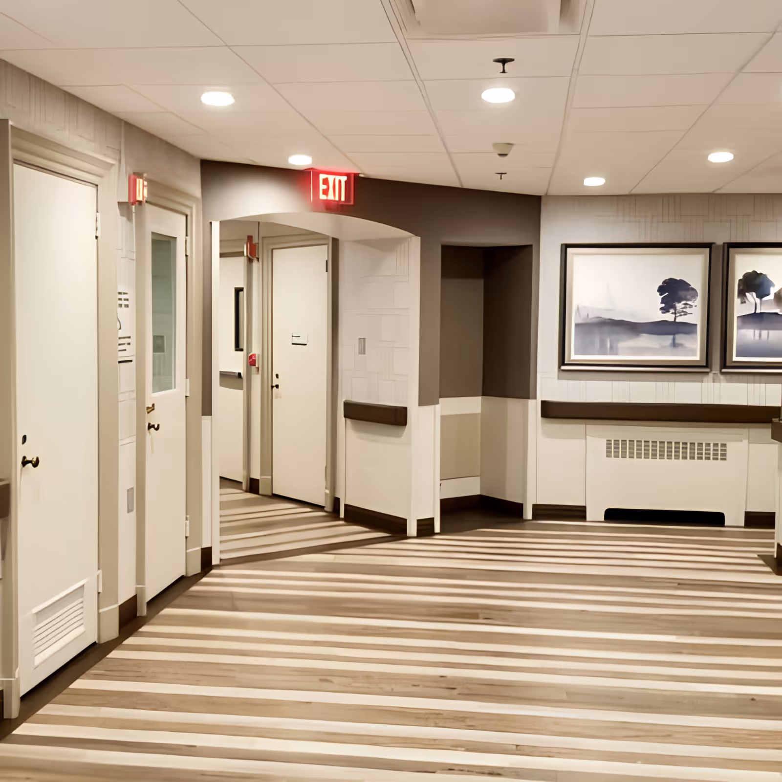 Interior hallway of a senior living facility with striped wooden flooring, white walls with brown accents, several closed doors, an illuminated red exit sign above an arched doorway, and two framed landscape paintings on the wall.