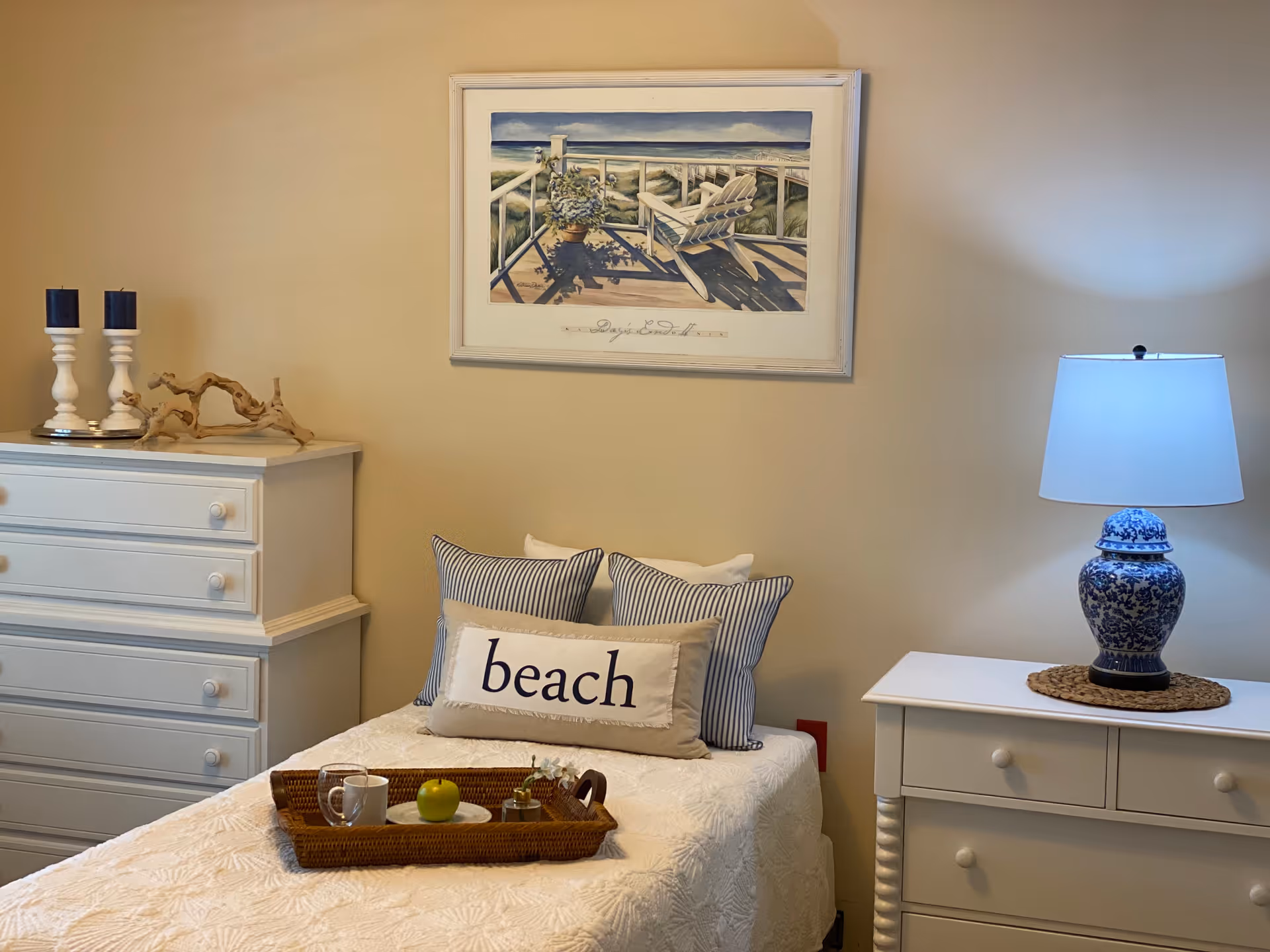 A cozy bedroom with a single bed covered in a white textured bedspread. The bed has three pillows, one with the word 'beach' printed on it. On the bed is a wicker tray holding a white mug, a green apple, and a small flower arrangement. To the left of the bed is a white dresser with two white candlesticks and a piece of driftwood on top. To the right is a white nightstand with a blue and white patterned lamp. Above the bed hangs a framed painting of a beach scene with a wooden deck, a chair, and a potted plant.