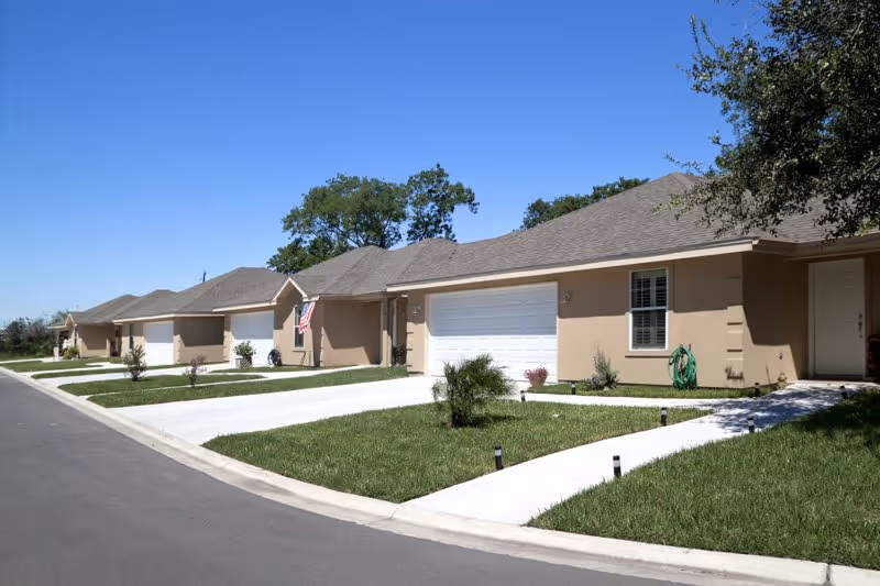 Single-story tan residential units with garages, driveways, and small lawns along a quiet street under a clear blue sky.