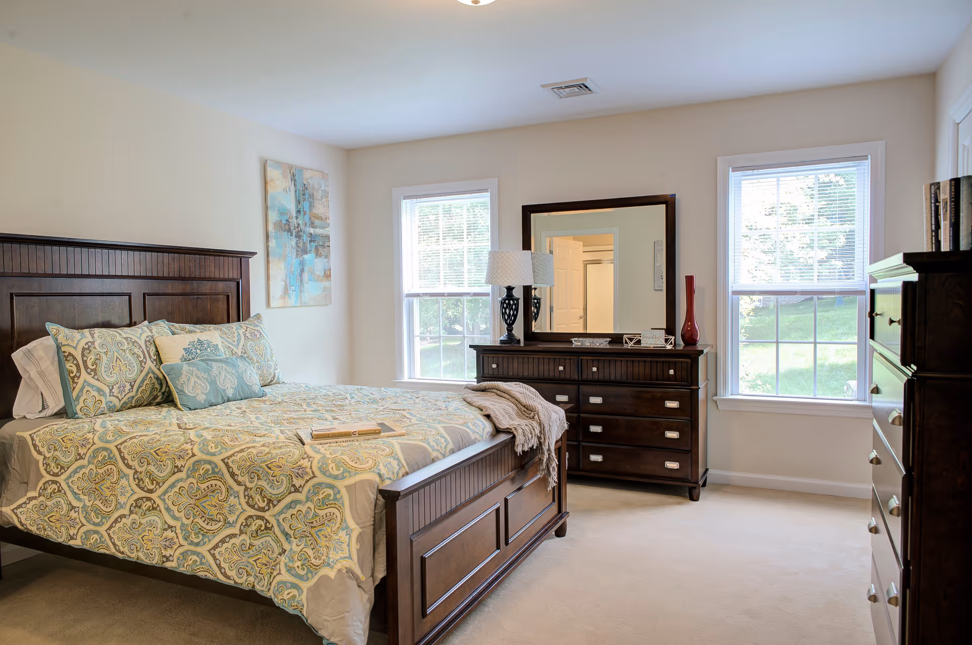 A bright bedroom with a large wooden bed featuring a patterned bedspread and multiple pillows. There are two windows letting in natural light, a wooden dresser with a mirror, a table lamp, and decorative items including a red vase. The walls are light-colored and there is a piece of abstract art hanging on one wall.