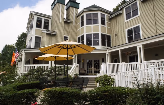 Exterior view of a multi-story senior living facility with beige siding and multiple windows. There is a patio area with black metal tables and chairs under large yellow umbrellas, surrounded by green bushes and plants. An American flag and another orange flag are visible on the left side near the building entrance.