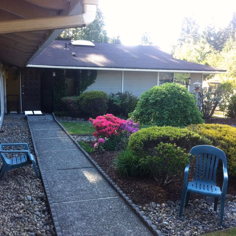 A paved walkway leading to the entrance of a single-story building with a garden on the right side featuring various bushes and colorful flowers. Two blue plastic chairs are placed on either side of the walkway, and the area is surrounded by trees and greenery.