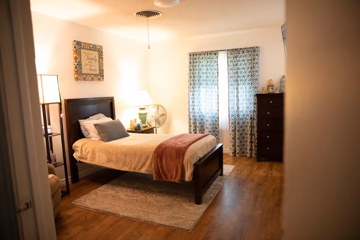 A cozy bedroom with a wooden bed frame, beige bedding, and a pink throw blanket. There is a bedside table with a lamp and a fan, a tall dresser, patterned curtains covering a window, and a framed wall decoration that reads 'The love of a Family makes this house a home.' The room has wooden flooring and a soft area rug under the bed.