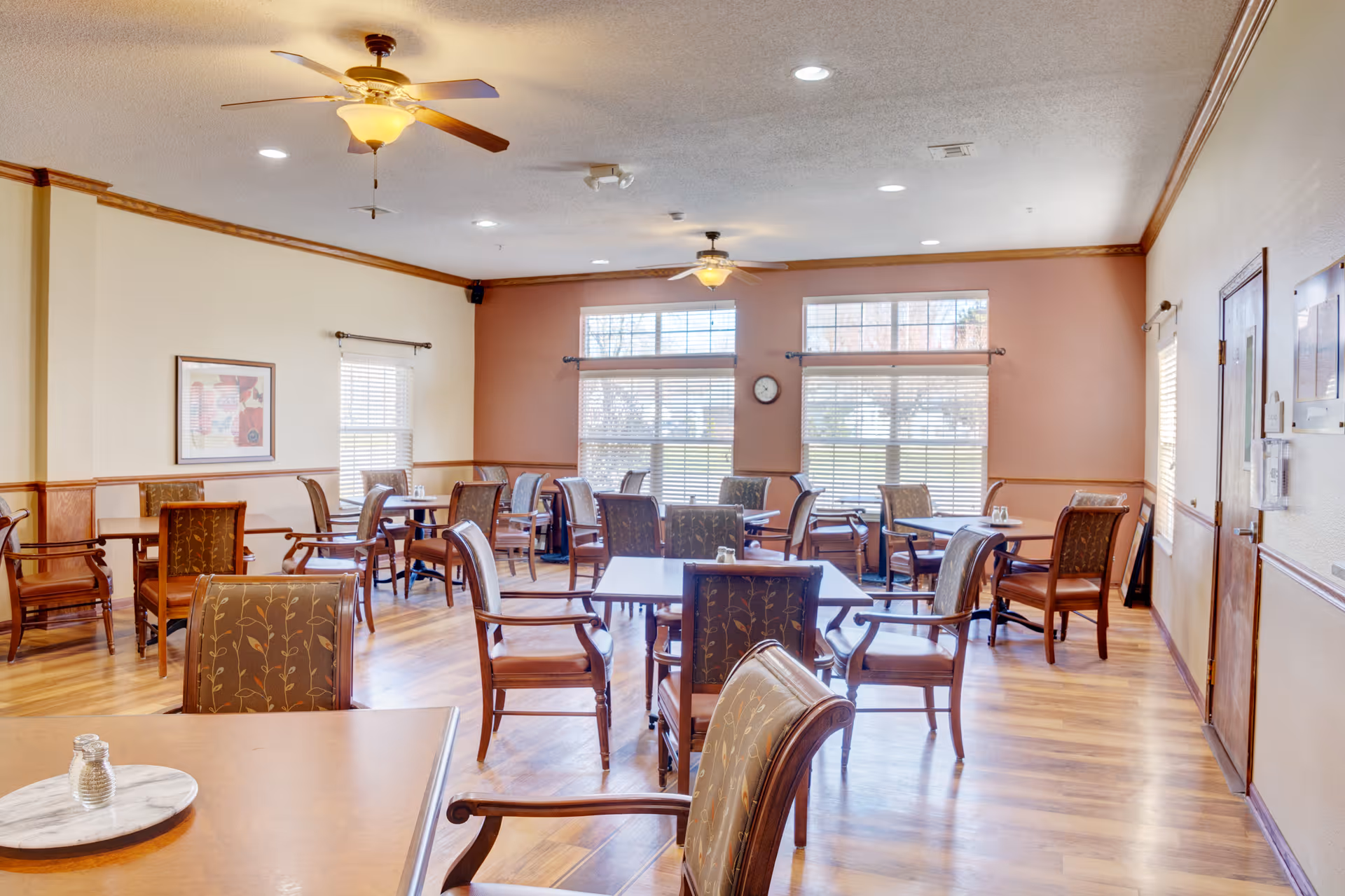 Bright communal dining room with multiple wooden tables and upholstered chairs, ceiling fans, and large windows.