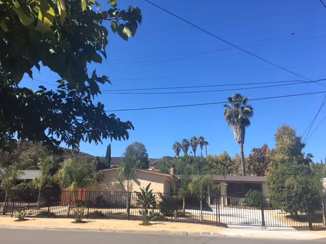 Exterior view of a single-story building surrounded by a black metal fence with various trees and plants in front, including palm trees, under a clear blue sky with visible power lines.