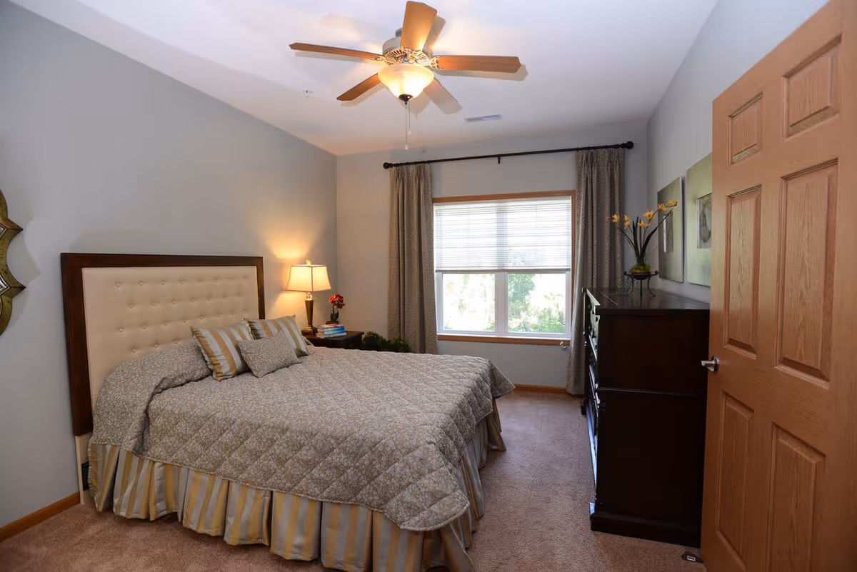 Cozy furnished bedroom with a bed and upholstered headboard, nightstand and lamp, dresser, window and ceiling fan.