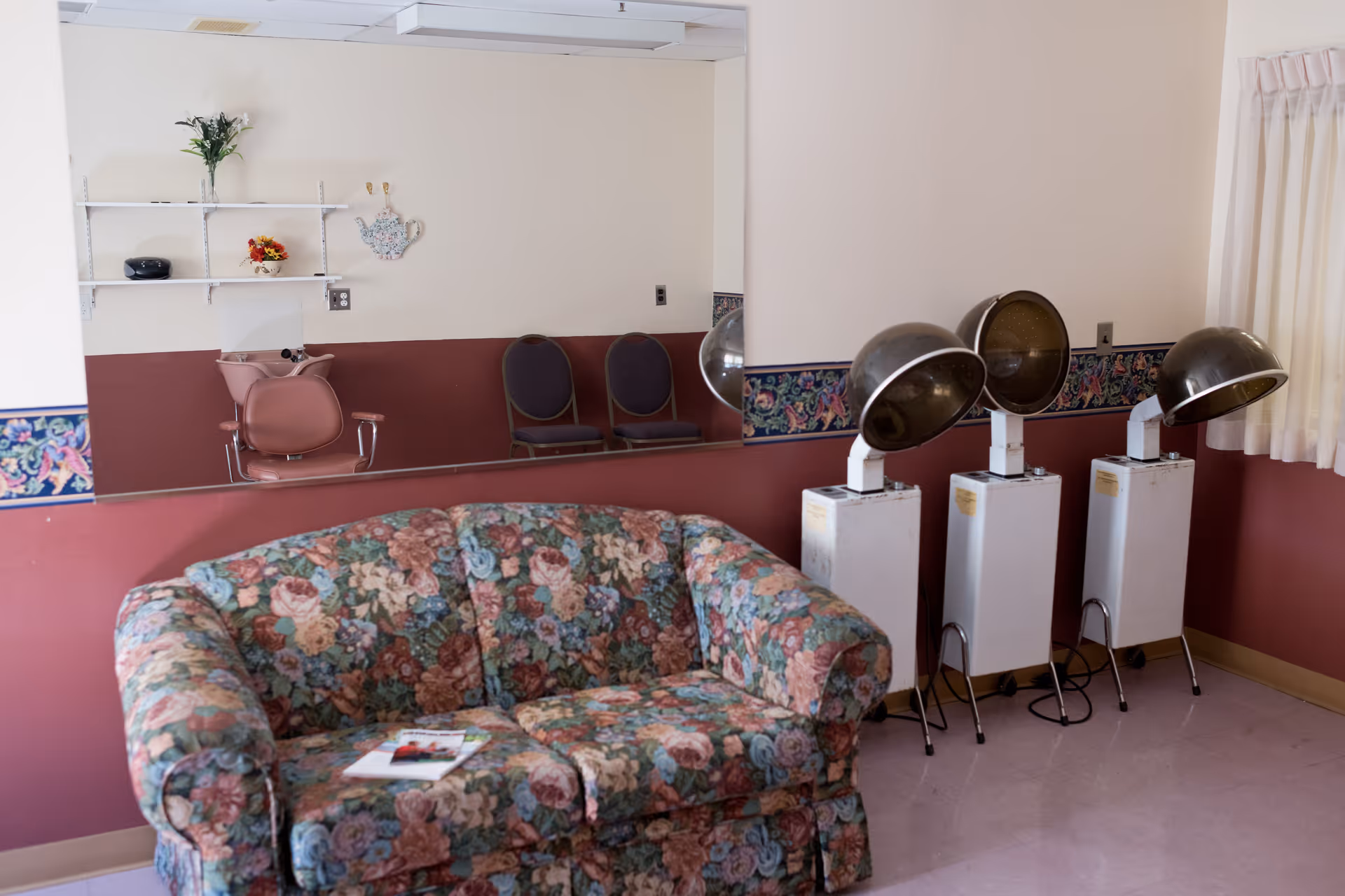 Floral-pattern sofa in a salon-style room with three hooded hair dryers, chairs, a large mirror and shelving.