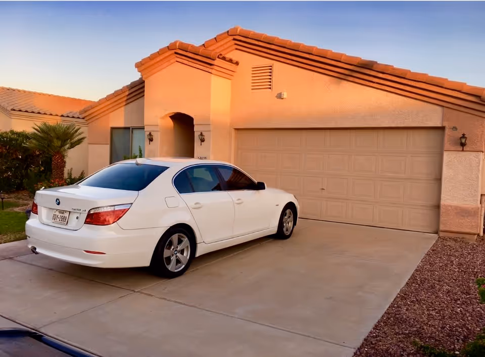 A white sedan car parked in the driveway of a single-story house with a beige exterior and a tiled roof during sunset.