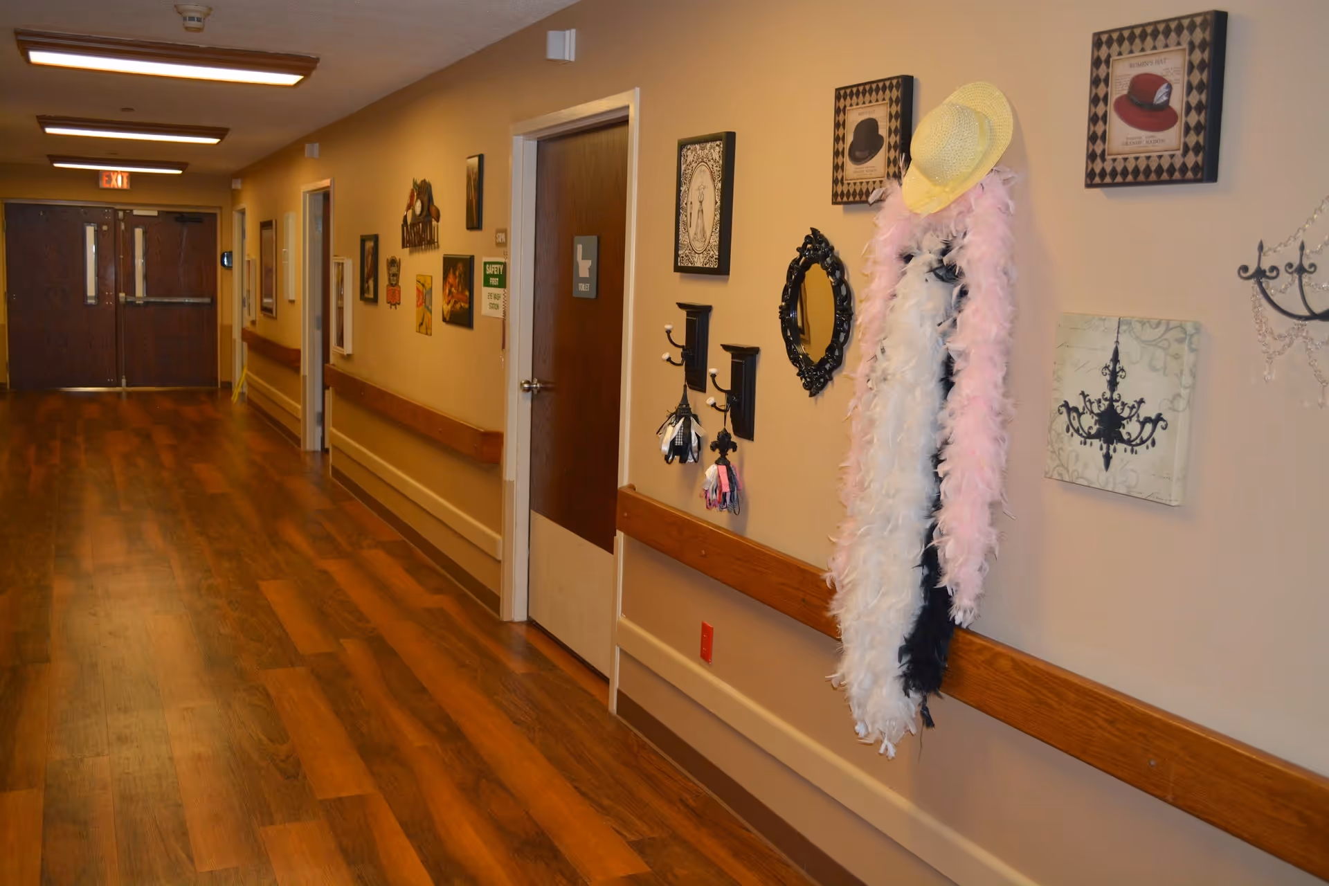 A hallway in Good Samaritan Home and Rehabilitative Center with wooden flooring and beige walls. The hallway features several doors, framed artwork, and decorative items including a yellow hat and feather boas hanging on the wall. There are handrails along the walls and ceiling lights illuminating the corridor.