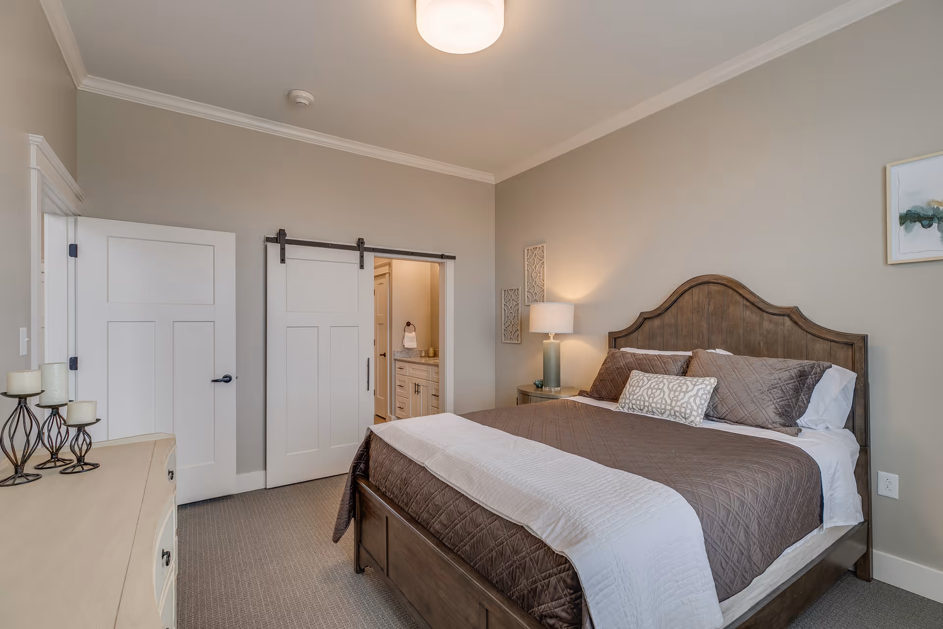 A neatly arranged bedroom with a wooden bed frame, brown quilted bedding, and multiple pillows. There is a nightstand with a lamp and decorative wall art above it. A sliding barn door leads to an adjoining bathroom, and a dresser with candles is visible on the left side of the room. The walls are painted beige and the carpet is light gray.