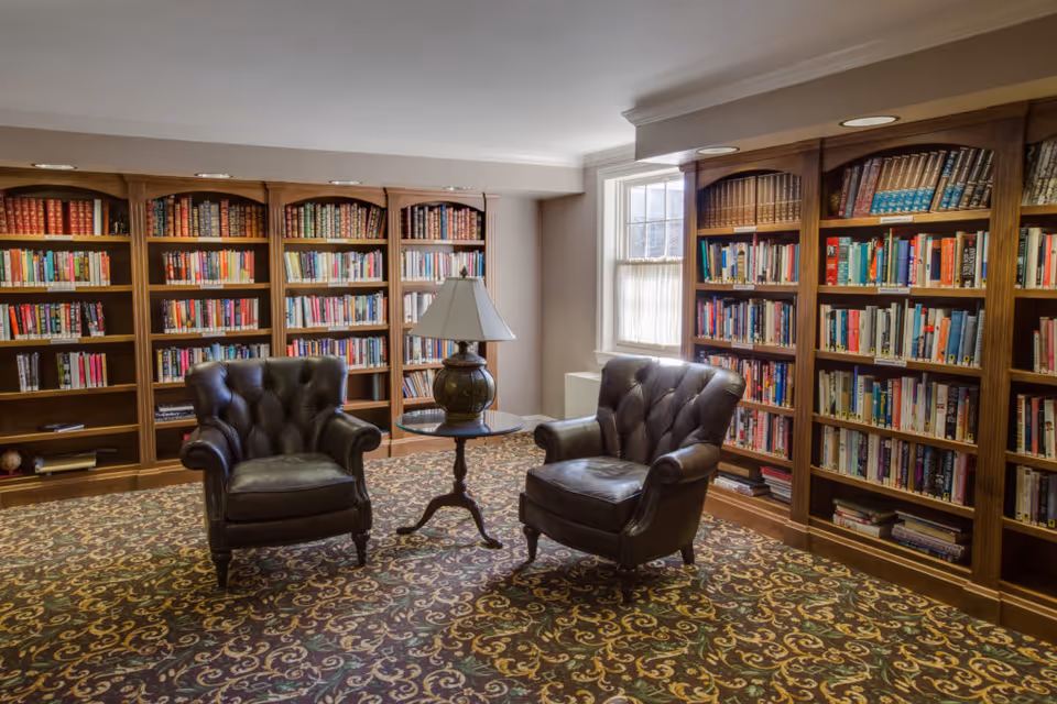 A cozy library-style sitting room with two leather armchairs, a small table and lamp, and bookshelves lining the walls.