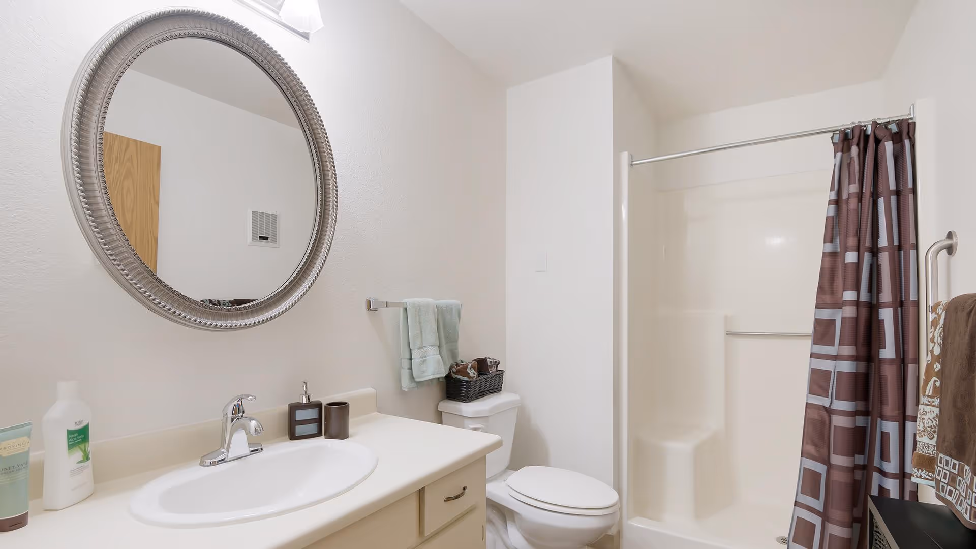 A clean and simple bathroom featuring a white sink with a silver faucet, a round decorative mirror above the sink, a toilet with a basket on top, and a shower with a brown and gray patterned curtain. Towels hang on racks near the sink and shower.