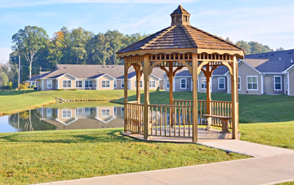 A wooden gazebo with a shingled roof situated on a grassy area near a small pond, with single-story residential buildings and trees in the background under a clear sky.