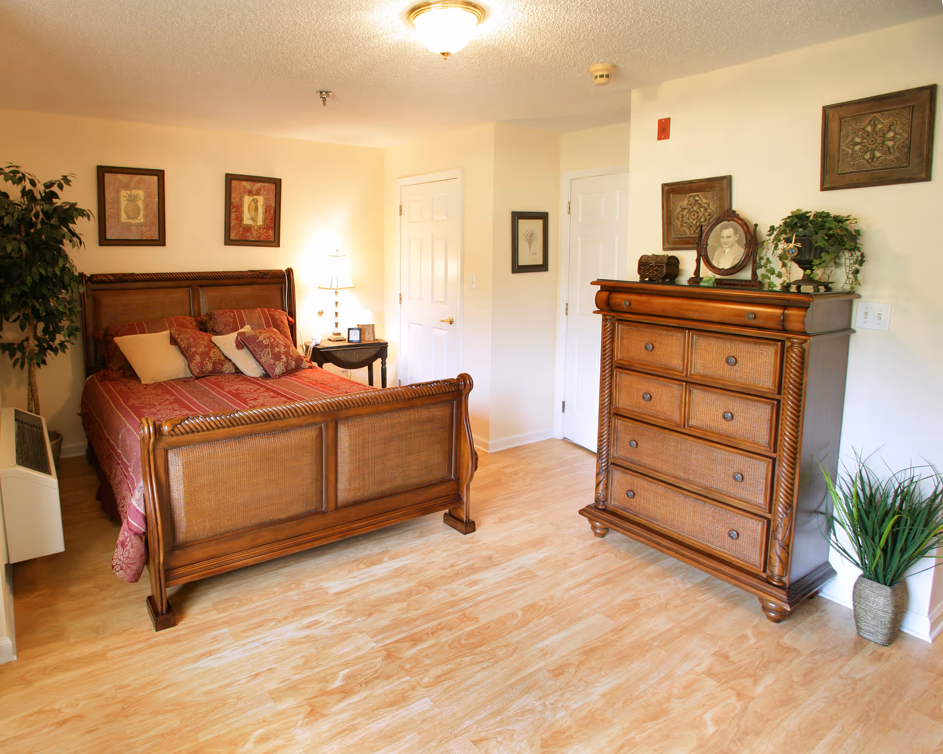 A cozy bedroom with a wooden bed frame and a red patterned bedspread, several pillows, a wooden dresser with decorative items on top, framed artwork on the walls, a small side table with a lamp, and potted plants in the corners.