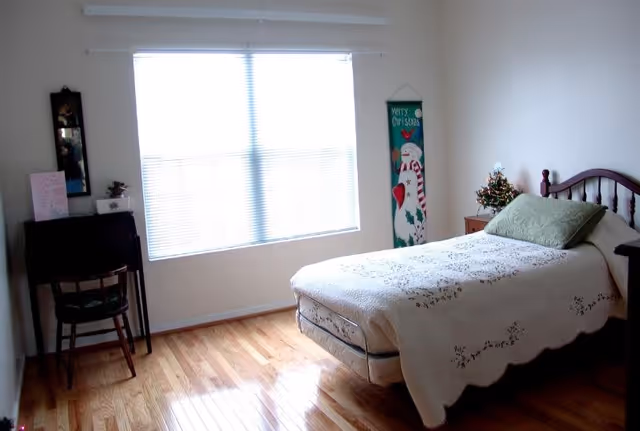 A bright bedroom with a single bed covered with a white quilt and a green pillow. There is a small wooden nightstand with a small decorated Christmas tree on it. A vertical Merry Christmas wall hanging with a snowman is on the wall next to the bed. A wooden chair and a small desk with a mirror above it are positioned near a large window with closed blinds. The floor is polished hardwood.