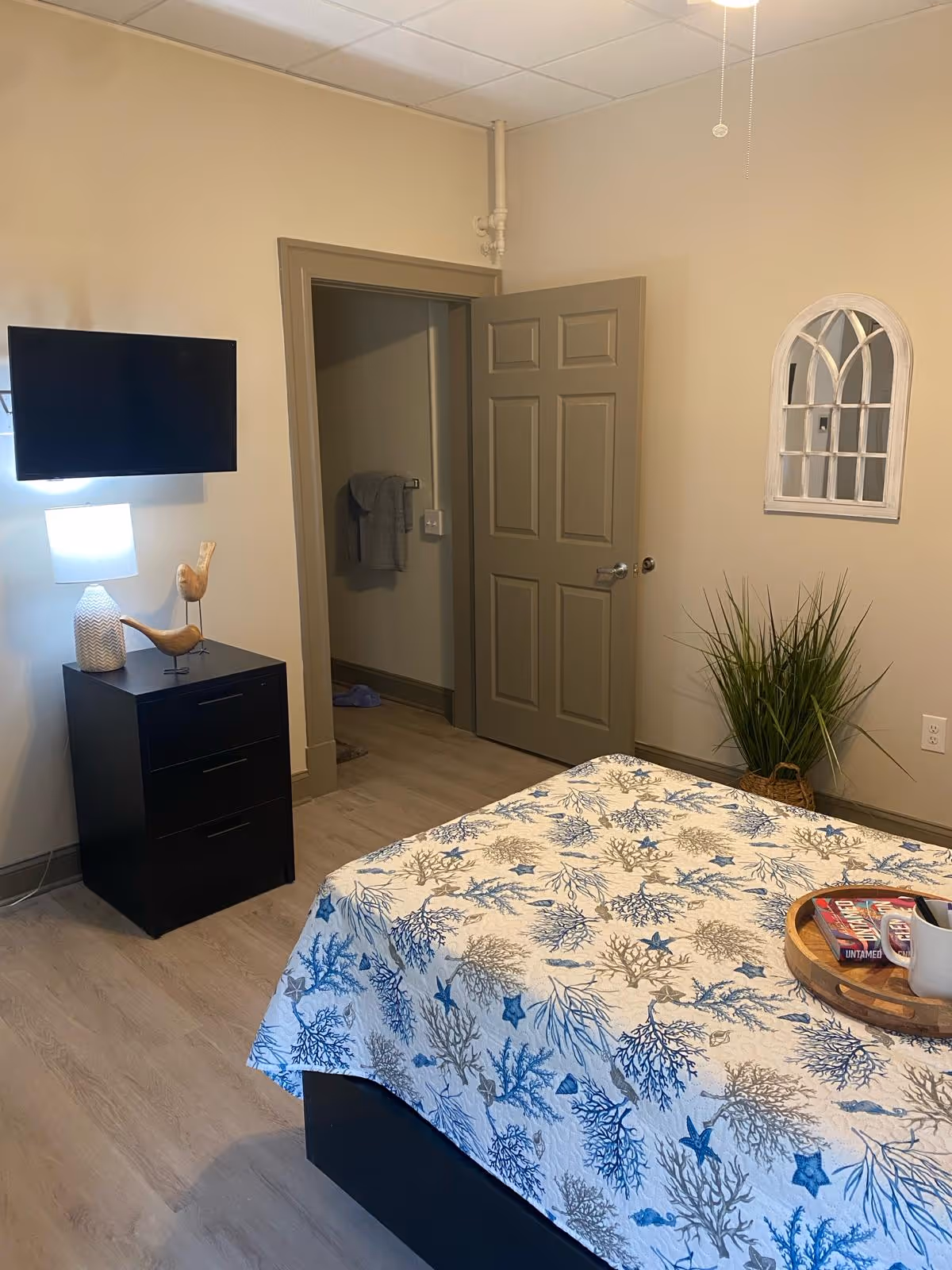 A bedroom in Ridgeway Village Assisted Living featuring a bed with a blue and beige sea-themed bedspread, a black nightstand with a lamp and decorative wooden birds, a wall-mounted TV, an open door leading to another room, a potted plant in the corner, and a decorative wall mirror shaped like a window.