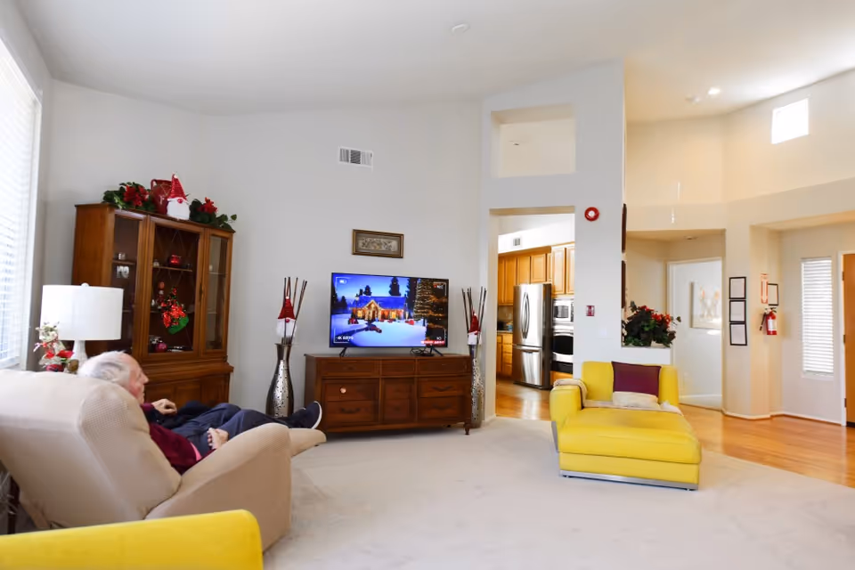 A bright living room with a beige armchair occupied by an elderly person, a yellow chaise lounge, a wooden cabinet with glass doors decorated with Christmas ornaments, and a flat-screen TV displaying a snowy holiday scene. The room has light-colored walls, carpeted floor, and an open view into a kitchen with wooden cabinets and stainless steel appliances.