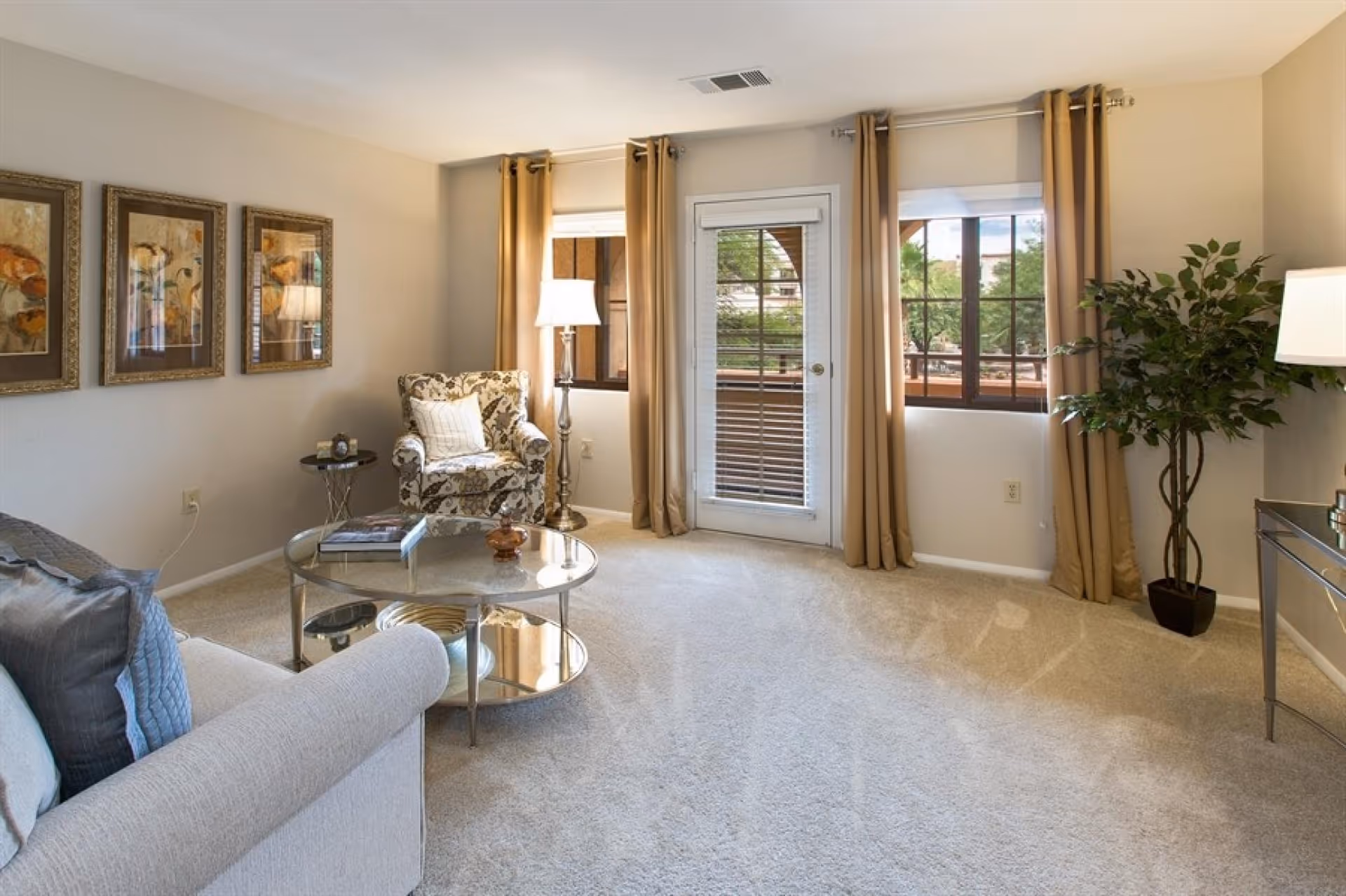 Bright living room with a sofa and patterned armchair around a glass coffee table, beige curtains framing a glass door and windows to an outdoor balcony.