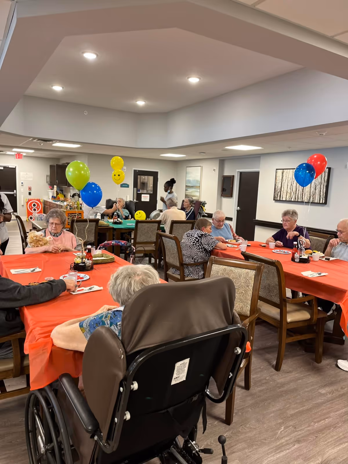 A group of elderly people sitting around tables covered with red tablecloths in a senior living facility dining area. Some are eating, while others are engaged in conversation. There are colorful balloons tied to the tables, and a caregiver is standing in the background. The room has bright lighting and neutral-colored walls with framed artwork.