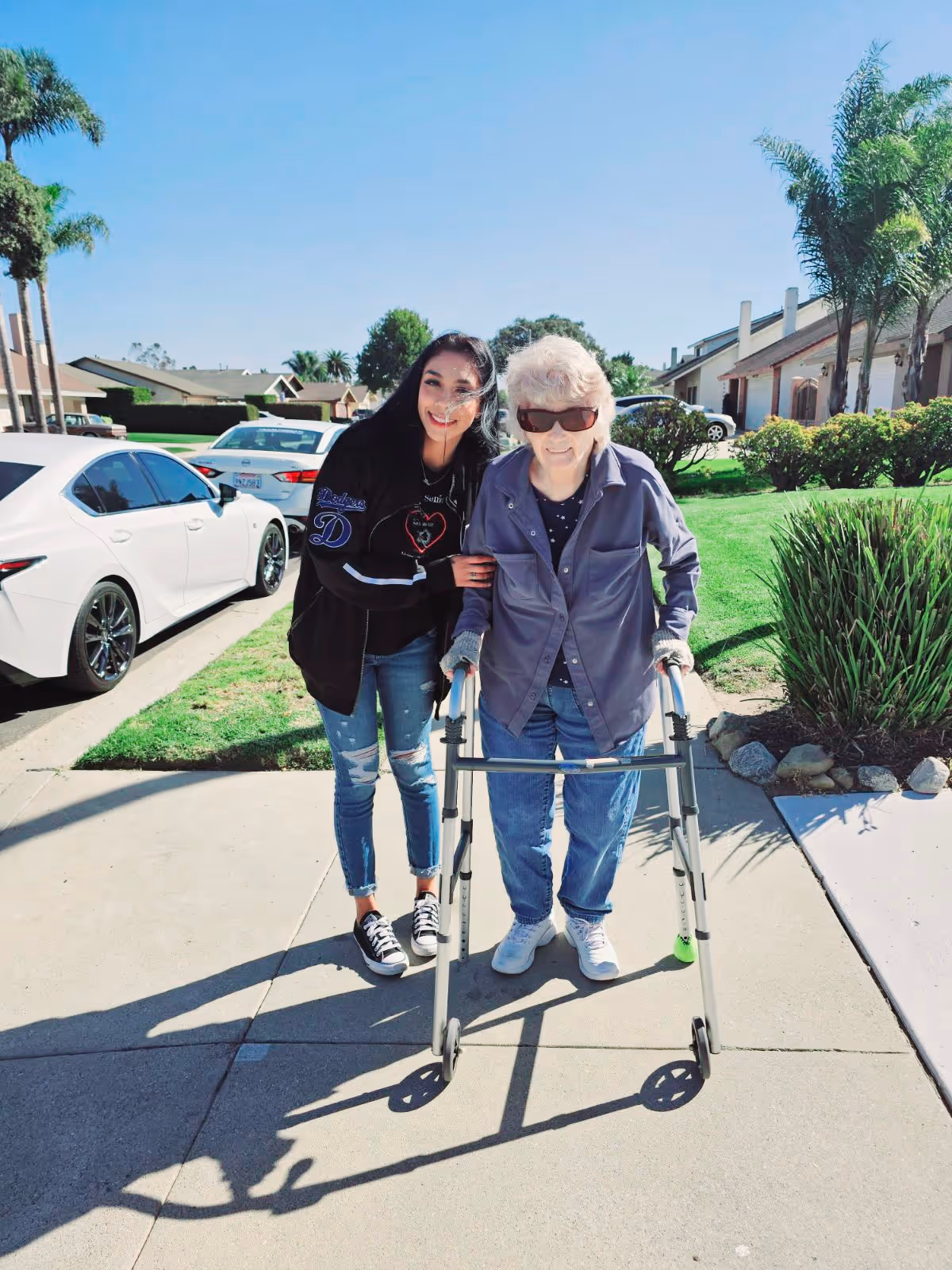 An elderly woman using a walker is standing on a sidewalk outside in a residential neighborhood, accompanied by a younger woman who is smiling and supporting her. There are cars parked along the street and houses with green lawns and palm trees in the background under a clear blue sky.