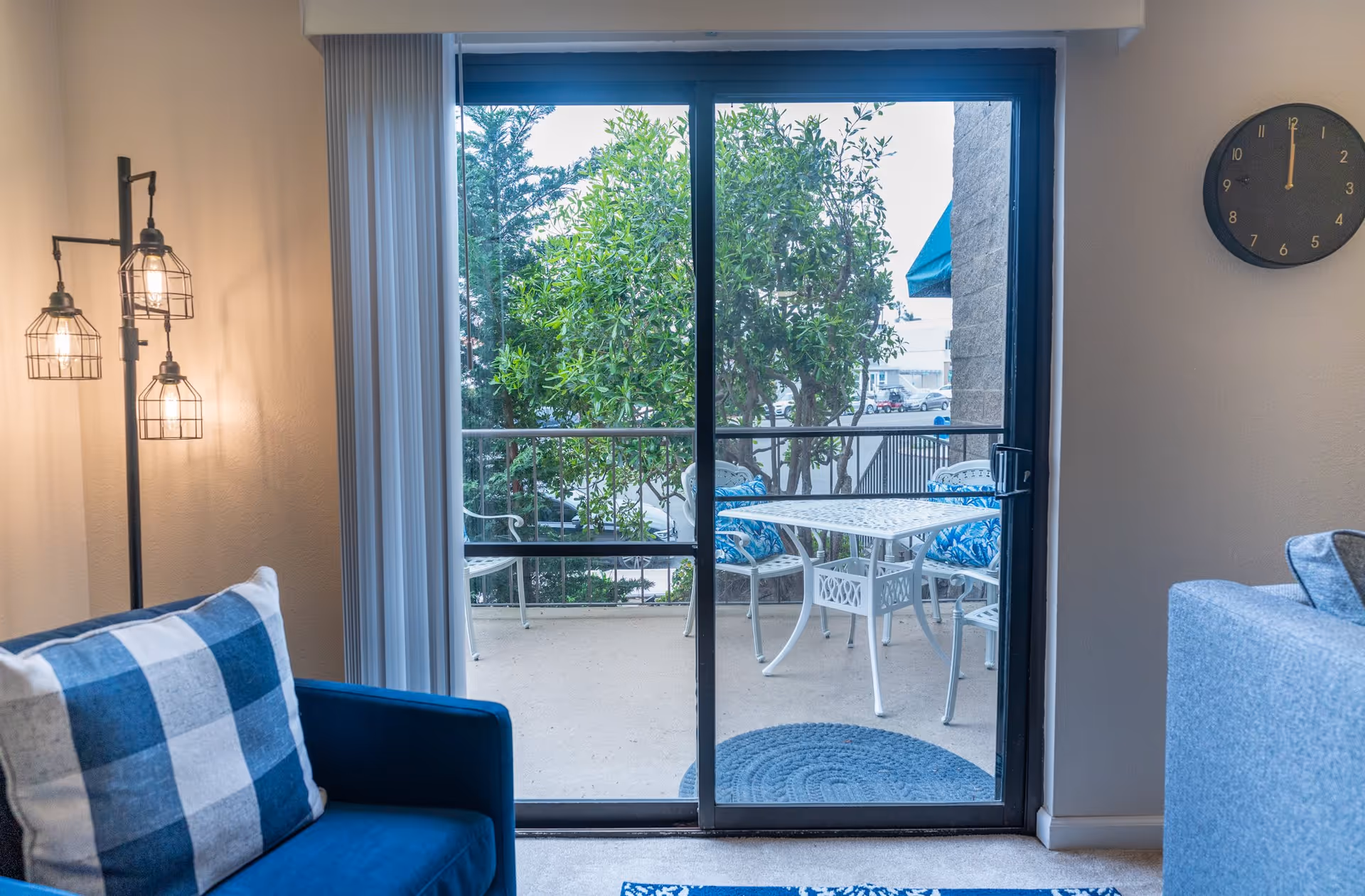 A cozy living area featuring a blue armchair and a glass door leading to a balcony with a table and chairs, surrounded by greenery.