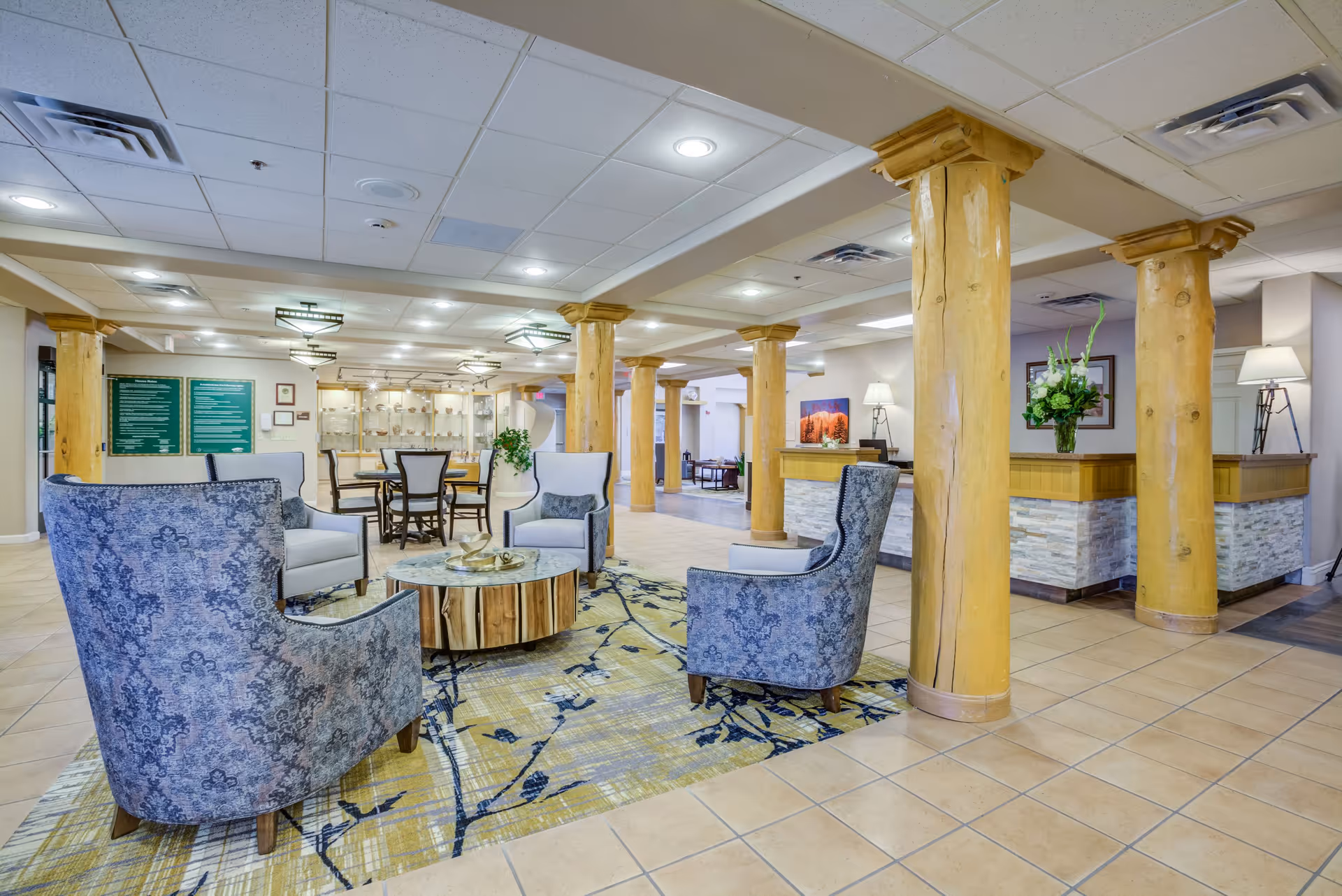 A spacious and well-lit senior living community common area featuring wooden pillars, a seating arrangement with blue patterned armchairs around a round wooden coffee table on a decorative rug, and a reception desk with a flower arrangement. The background includes additional seating and wall decorations.