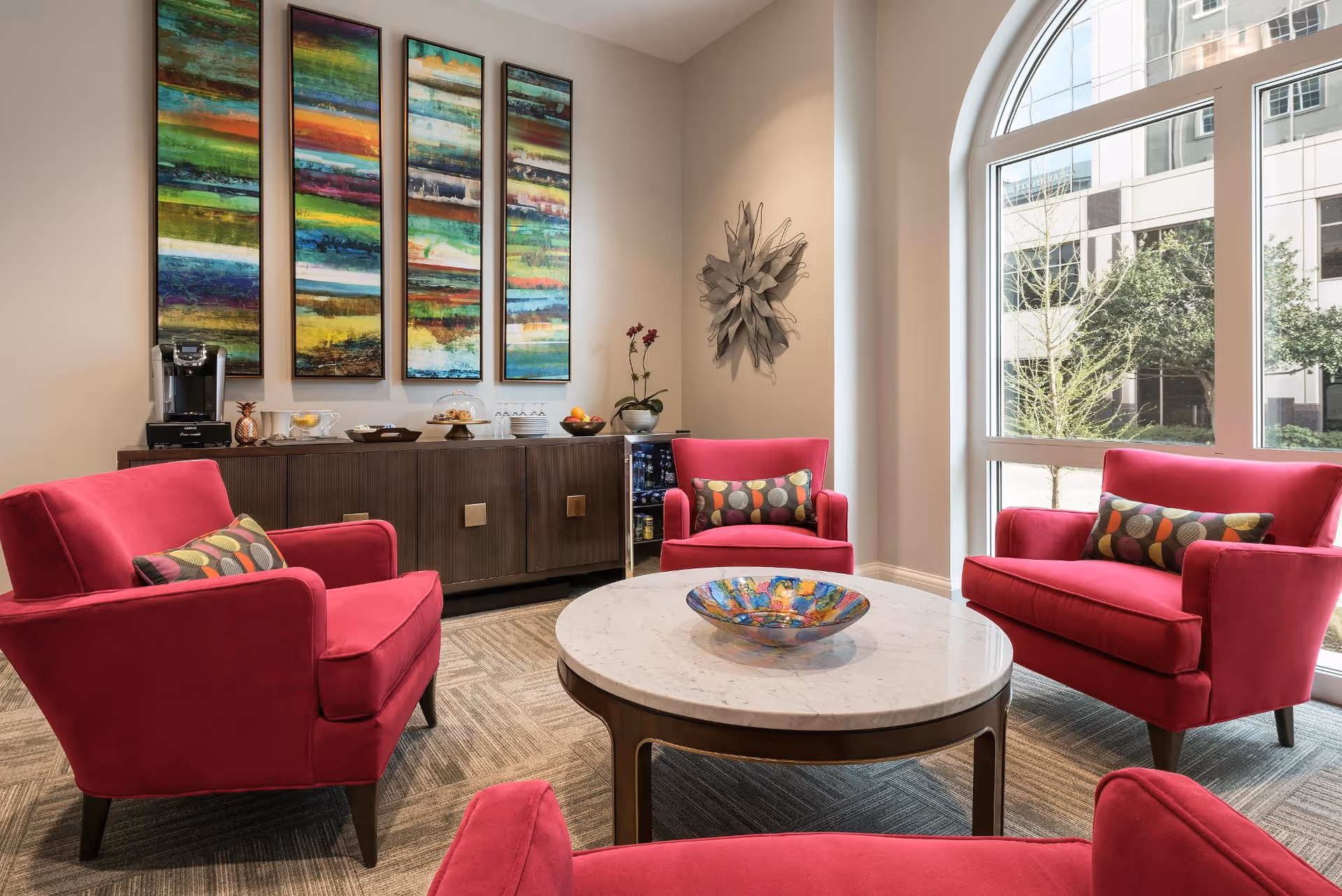 Bright seating area with four pink armchairs around a round marble coffee table in a lounge with a large arched window and colorful wall art.
