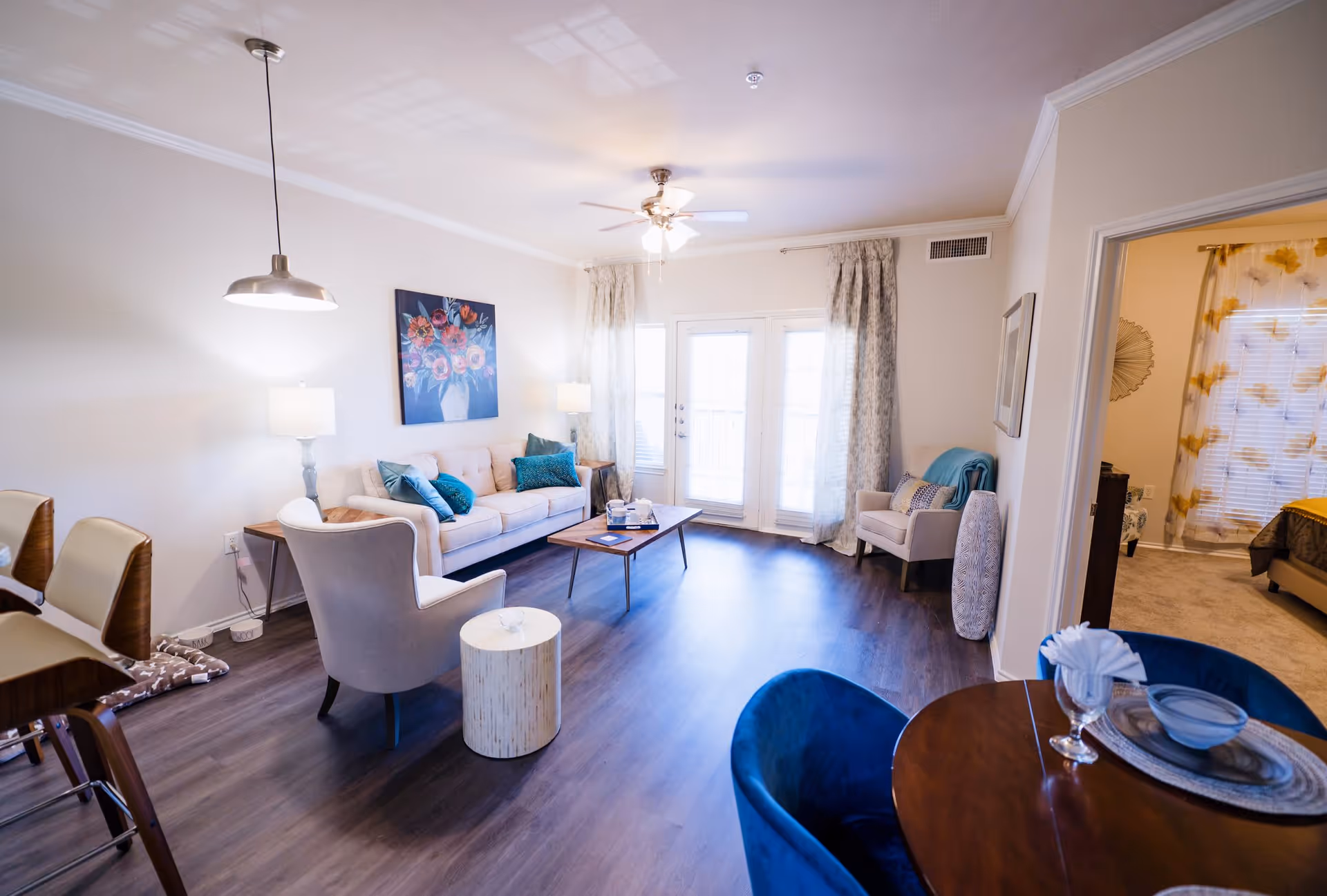 A bright and modern living room with a beige sofa adorned with teal pillows, two armchairs, a wooden coffee table, and a side table with a lamp. The room has dark wood flooring, a ceiling fan with lights, and large French doors with sheer curtains letting in natural light. Adjacent to the living room is a dining area with a round wooden table and blue velvet chairs. A bedroom with floral curtains is visible through an open doorway.