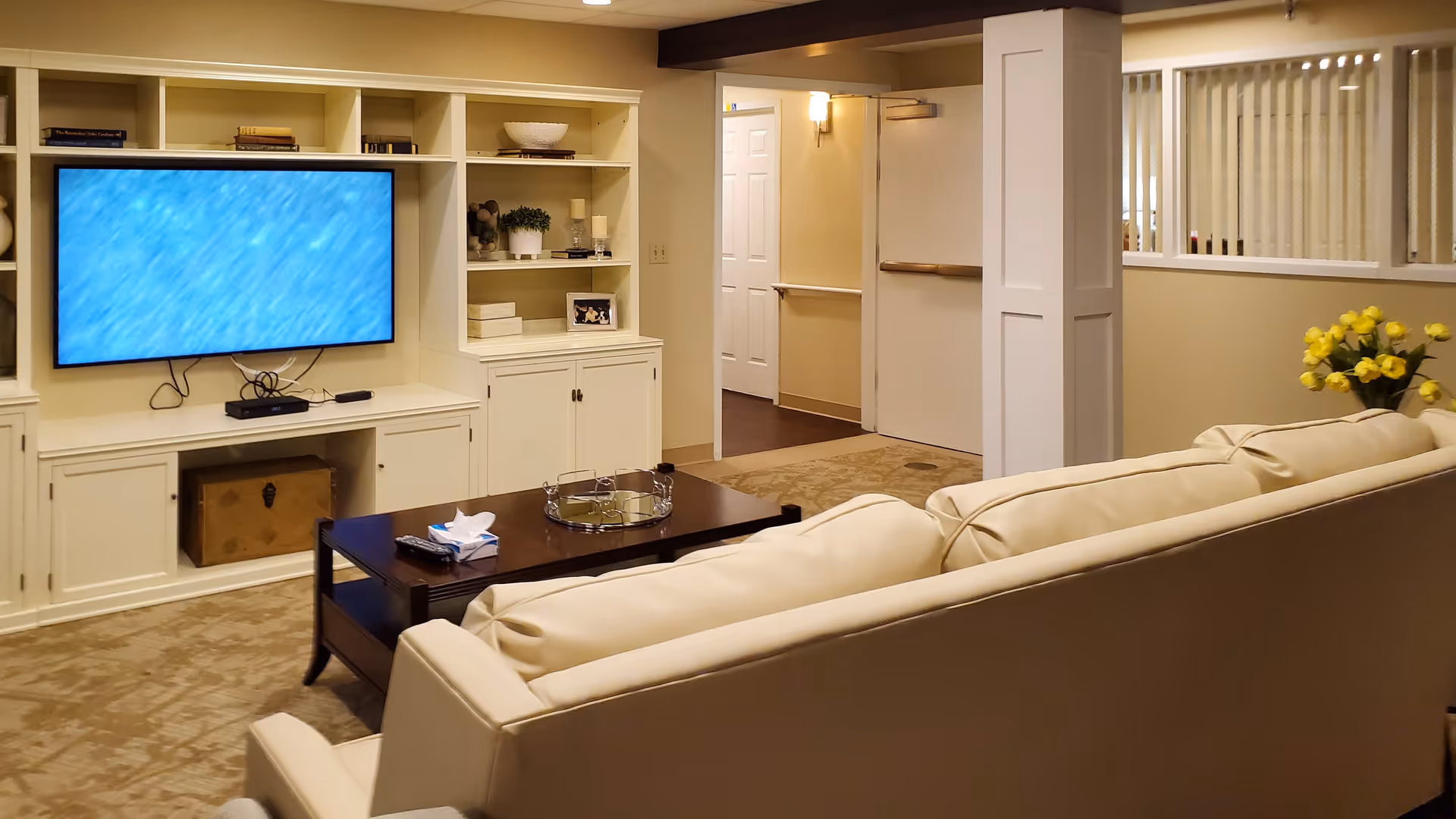 A cozy living room area with a beige sofa facing a wall-mounted flat-screen TV. The TV is set within a white built-in shelving unit that holds decorative items such as books, plants, and candles. A dark wooden coffee table in front of the sofa holds a tissue box, a remote control, and a decorative tray. The room has beige walls and carpet, with a doorway leading to a hallway. A vase with yellow flowers is placed near the sofa.