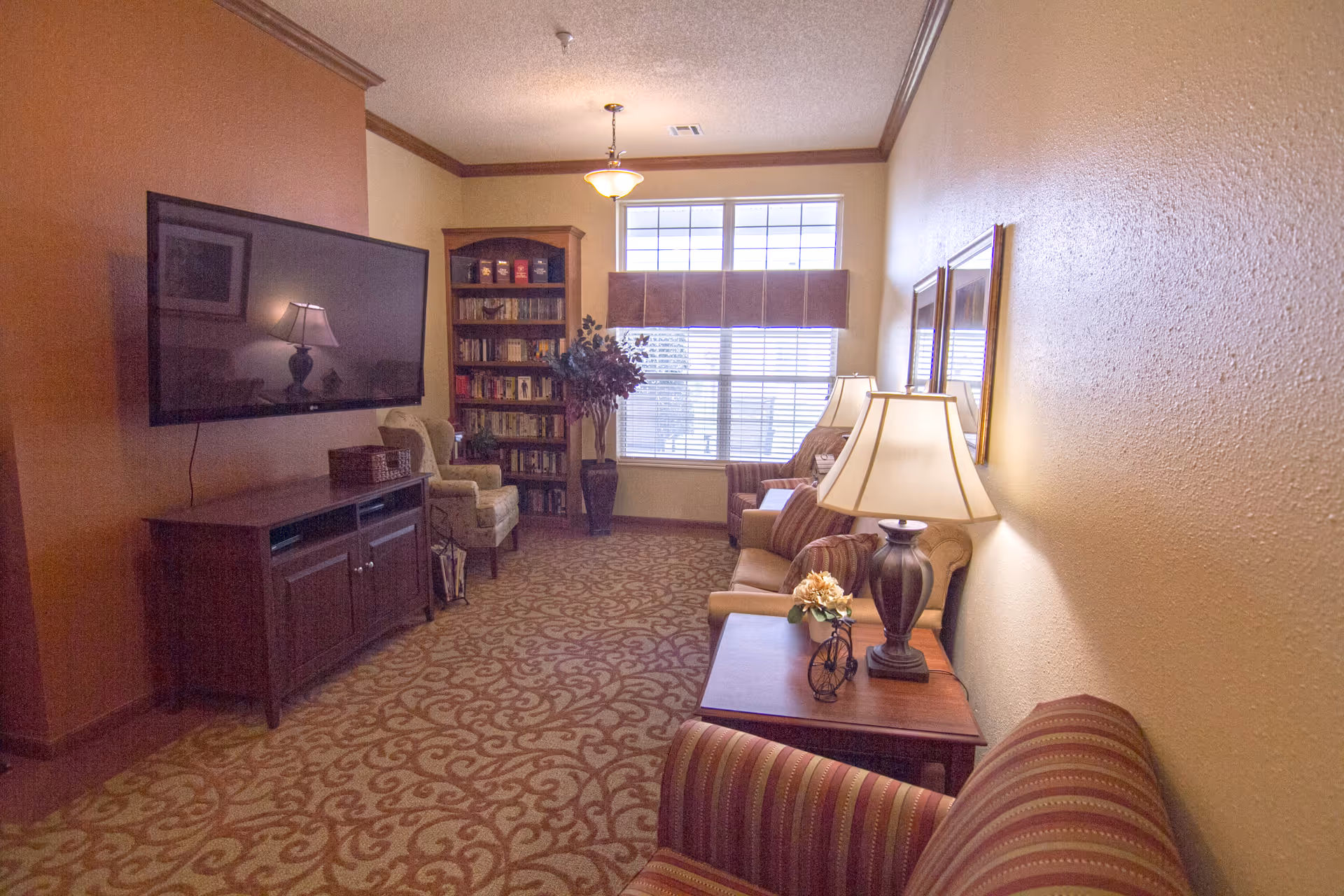 A cozy living room with patterned carpet, a large flat-screen TV mounted on a burnt orange accent wall, a wooden TV stand, a bookshelf filled with books, two armchairs, a sofa with striped cushions, two table lamps on wooden side tables, and a large window with blinds and a valance letting in natural light.