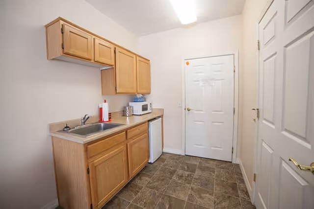 Small kitchenette with wooden cabinets, a sink, microwave, mini fridge and a closed door.