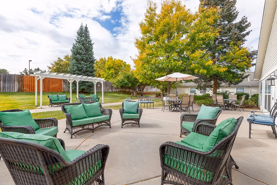 Outdoor patio area with multiple wicker chairs and sofas featuring green cushions arranged in a circle on a concrete surface. In the background, there are tables with umbrellas, a white pergola, green grass, trees with autumn foliage, and a wooden fence.