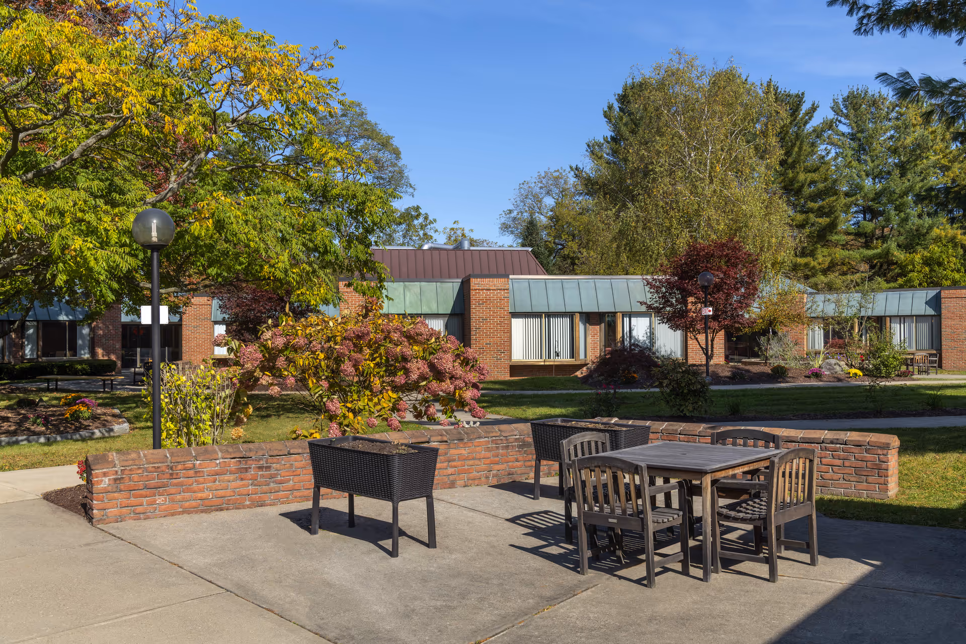 Outdoor patio area at Elderwood at Waverly featuring a concrete surface with a table and four chairs, surrounded by a low brick wall. In the background, there are well-maintained gardens with various trees and shrubs, and a single-story brick building with large windows under a clear blue sky.